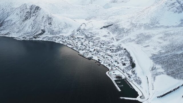 Aerial view of the small village of Fjordgard nestled beside dark waters and snow-blanketed mountains near Segla, Fjordgard, Troms, Norway.