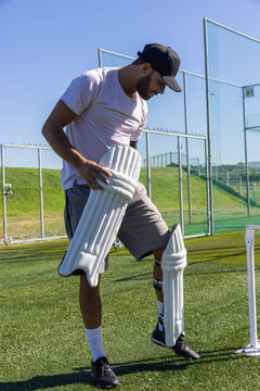 Batting pads are being fastened on artificial turf near white stumps and netted practice cages