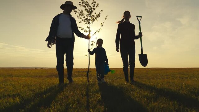 A family of three is walking through a field with a tree in the foreground. The man is holding a shovel and the woman is holding a tree sapling