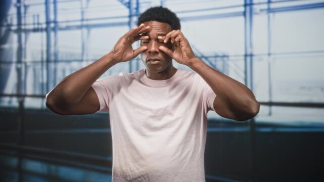 Black man pulls down lower eyelids with fingers and rolls eyes in airport terminal wearing pink tshirt and looking upward; tired travel delay.