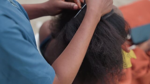 Closeup black stylist creating neat rows with tail comb, precise parting and threading motions, focused professional technique in outdoor market setting, rhythmic handwork and careful tension control