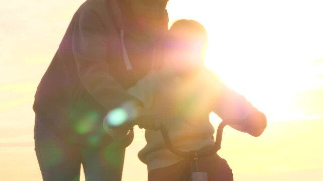 Silhouettes of child and mother, riding bike at dawn, sunset. Parent in field teaching educating girl skill of riding bicycle. Glare from sunrise sun, family summer fun. Mom training instructing kid.