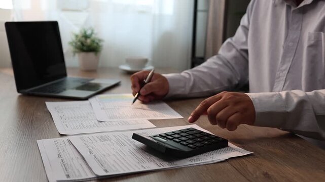 Close-up of a person calculating numbers with a calculator while filling out documents at a desk with a laptop in a home or office setting, suitable for finance, accounting, and paperwork tasks.