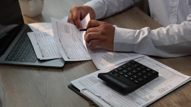 Close-up of a person pressing a calculator while holding a pen and reviewing financial documents on a desk with a laptop and coffee cup, suitable for accounting, budgeting, tax filing, expense trackin