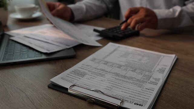 Close-up of a person reviewing and writing on financial forms clipped on a clipboard with a pen while holding documents at a desk with a laptop and calculator, suitable for budgeting, tax filing, and 
