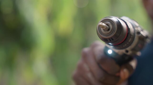 Closeup showing carpenter holding drill, outdoor bokeh background, steady grip on power drill, LED light on, metal chuck and worn bit visible, atmosphere of focused craft and preparatory carpentry