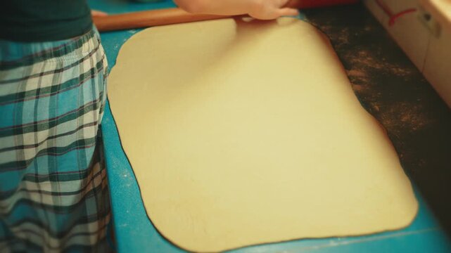 Close up shot of teenage hands using a wooden rolling pin to flatten raw yeast dough on a blue counter. Authentic home baking process for sweet pastry and dessert.