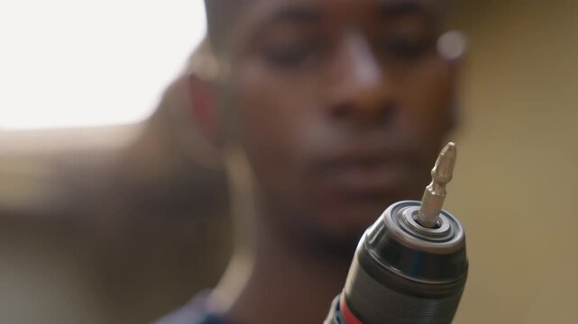 Black apprentice carpenter inspects drill closeup, focused young black man in blurred workshop background, power drill held steady with metal bit glinting, concentrated expression, carpentry training