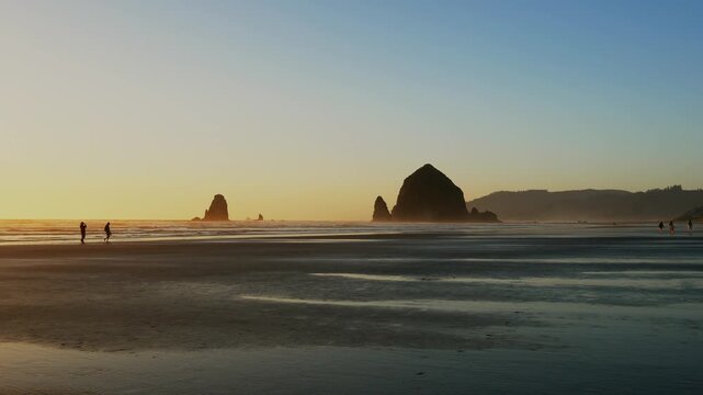 Haystack Rock and Needles at Cannon Beach during sunset with people walking along wet sand and gentle waves creating scenic Oregon coastal landscape with reflections