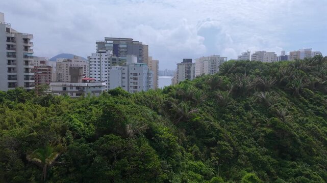 Praia das Asturias beach in Guaruja, Sao Paulo, Brazil, revealed through coastal Atlantic Forest with dense high-rise buildings and toweres with ocean view, urban coastal landscape, drone push in