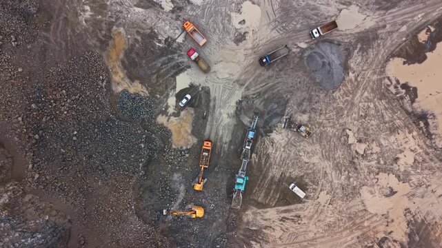 Heavy machinery and trucks work at a granite quarry site. Excavators dig into the ground, and trucks transport materials. The site shows signs of ongoing development and extraction.