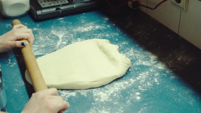 Close up shot of teenage hands using a wooden rolling pin to flatten raw yeast dough on a blue counter. Authentic home baking process for sweet pastry and dessert.