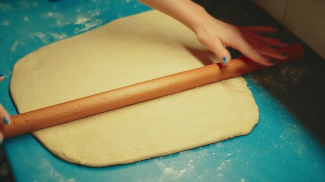Close up shot of teenage hands using a wooden rolling pin to flatten raw yeast dough on a blue counter. Authentic home baking process for sweet pastry and dessert.