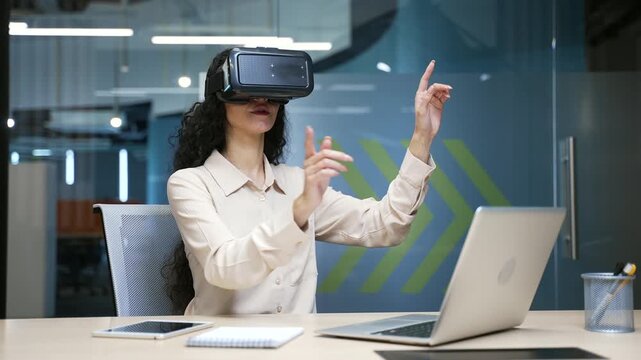 Confident businesswoman works using VR glasses in virtual reality simulator while sitting in a business office. The female uses gestures to control computer programs and flips through virtual pages