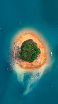 Aerial view of a small circular island with lush green trees and orange soil surrounded by turquoise water and a lone kayak with a person in Khao Sok National Park, Surat Thani, Thailand.