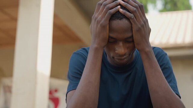 Distressed Black carpenter holding head outdoors at worksite, porch columns and roofline in background, visible sweat and tense posture, moment of worry over project setback, emotional candid portrait