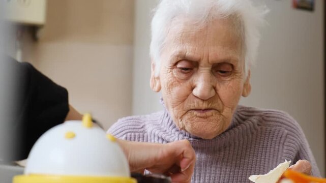 Very elderly senior woman eating at table while someone hand serves extra food into her plate. Elderly woman receiving care and love smiles gratefully. Care for lonely elderly and charity concept