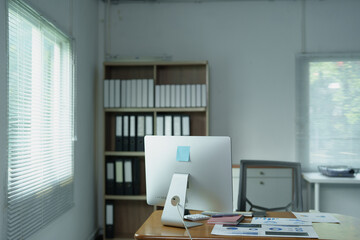 Modern office workspace with computer monitor on wooden desk, paperwork, calculator and keyboard, bright window light, organized bookshelf of binders and files for professional use © MINAE