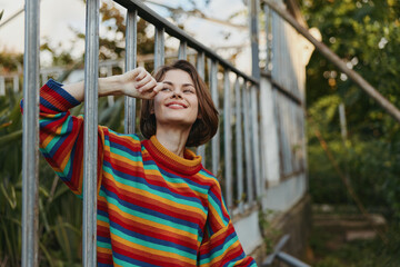 Fototapeta premium Woman in a striped sweater leaning against a greenhouse metal rail, smiling and touching her face, short hair, casual outfit in a sunlit outdoor garden lifestyle moment.