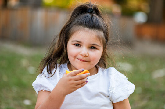 Portrait of young girl eating a mango slice while standing in park with greenery behind her