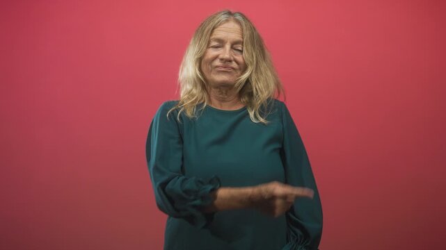 Senior woman points finger to side and touches shoulder in studio with pink backdrop; skepticism reflection.
