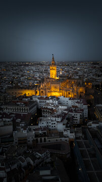 Aerial view of the illuminated Seville Cathedral and Giralda tower overlooking the historic city center at dusk in Seville, Andalusia, Spain.