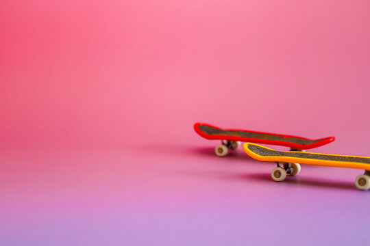 ​A pair of colorful finger skateboards, one red and one yellow, positioned together on a pink-to-purple gradient background. These toy miniatures perfectly capture the spirit of street sports and yout