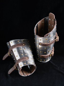 pair of metal bracers with leather straps and brass rivets designed for medieval armor. Displayed on black surface these items embody historical craftsmanship for reenactments or collectibles.