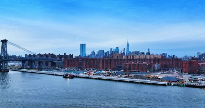 View on the waterfront of the East River with construction in progress. Approaching the Williamsburg Bridge and city skyline at dusk.