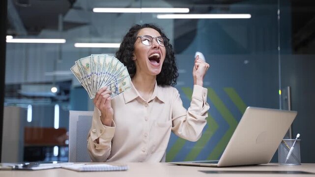 Happy excited businesswoman holding fan of cash money dollar bills sitting in office looking at camera. Joyful professional woman celebrates financial success by throwing money in air, falling dollars