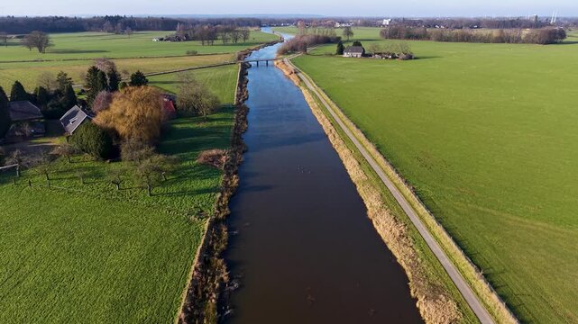 Aerial view of a rural landscape with a canal curving through green fields, two small road bridges crossing it, and a nearby house with outbuildings set among open farmland.