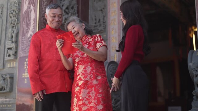 Asian grandparents receiving red envelope from young woman at chinese temple, joyful moment of giving during lunar new year celebration, showing love, family connection and festive cultural tradition