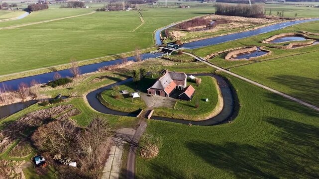 High angle drone view of a straight Dutch canal cutting through vibrant green pastures, featuring a traditional farmhouse, a small water control gate, and a parallel cycling path.