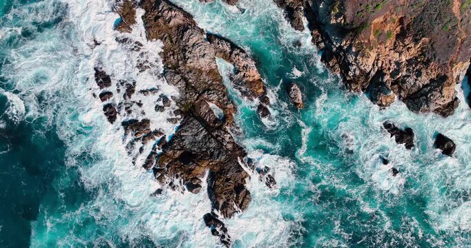 Aerial view of turquoise ocean waves crashing against jagged rocks. Top down drone shot of Pacific Ocean surf hitting dark coastal rock formations in California.