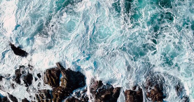 Abstract aerial texture of ocean surf hitting rocky shore. High angle shot of white water and sea foam swirling around dark coastal stones.