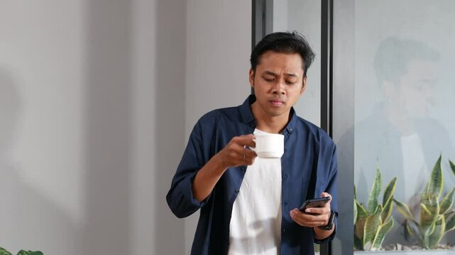 Confident Asian man coming to window at home, drinking hot tea or coffee and scrolling screen of her smartphone, checking messengers and social media