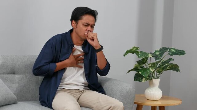A young Asian man coughing indoors, looking unwell, denoting sickness or allergies while sitting on couch at home