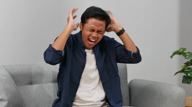 Angry dissatisfied Asian man sitting on sofa in living room at home. Frustrated male screaming, talks about problems that need to be solved