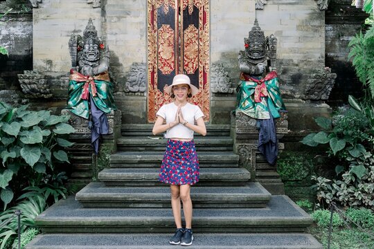 Woman in white shirt and colorful skirt standing in prayer on stone steps in front of ornate temple door with guardian statues
