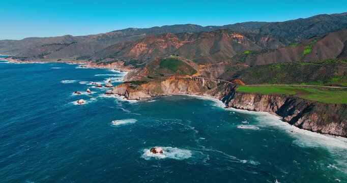 Bixby Creek Bridge and rugged California coastline at daytime. Wide aerial view of historic concrete arch bridge crossing a deep canyon along the Pacific coast.
