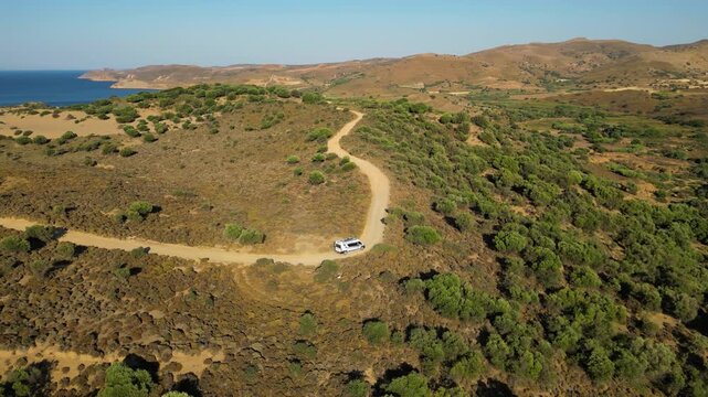 Aerial view of a white camper van traveling along a winding dirt road through the rugged, golden landscape of Limnos Island, Greece, highlighting the freedom of off-grid exploration.