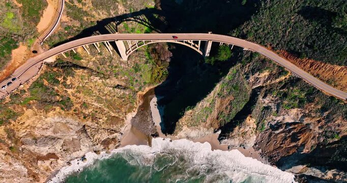 Bird's eye view of Bixby Creek Bridge with red car. Top down aerial perspective of an arched concrete bridge spanning a canyon over a beach in Big Sur, California.