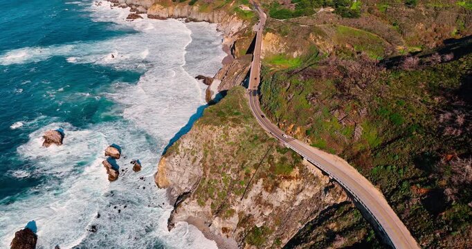 Aerial view of Pacific Coast Highway winding along Big Sur cliffs. High angle shot of Highway 1 winding through green coastal mountains next to turquoise ocean waves in California.