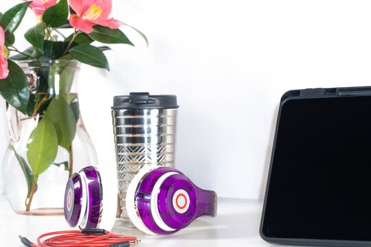Top view of a workspace with black tablet, purple headphones, pink flowers and metal cup on white background. Composition with space for text.