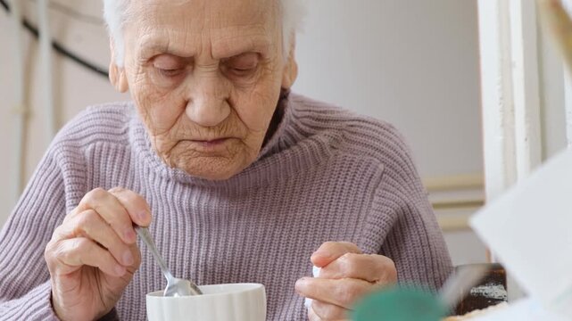 Gray haired wrinkled senior woman slowly chewing food and using metal teaspoon to stir tea in cup. Feeding and care for elderly people. Kindness and charity concept