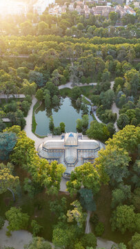 Aerial view of the Palacio de Cristal glass pavilion and its pond surrounded by lush green trees in Retiro Park Madrid, Community of Madrid, Spain.