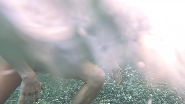 Woman swimming underwater, beautiful female in red swimsuit enjoying sea on pebble beach, emerging to surface with joy