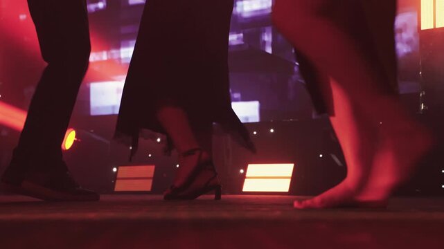 Closeup dancing feet on redlit stage, intimate couple steps as man in shoes and barefoot woman glide, warm spotlights and shimmering background panels create sensual club atmosphere