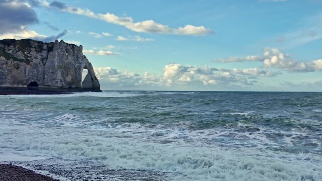 Aval cliffs with natural chalk arch and waves hitting pebble beach in Etretat, Normandy, France, dramatic coastal geology shaped by erosion along the English Channel, horizontal pan, wide shot