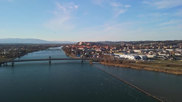 Ptuj, Slovenia, features its historic castle, medieval old town, red-roofed buildings, and bridges spanning the Drava River, with Ptujsko Jezero and surrounding hills visible in a daytime panorama.